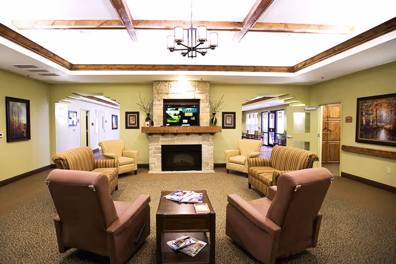 A cozy living room area in The Towne Care facility featuring a stone fireplace with a TV mounted above it, surrounded by four armchairs and two striped sofas arranged around a wooden coffee table with magazines. The room has light green walls, framed artwork, and a ceiling with wooden beams and a chandelier.