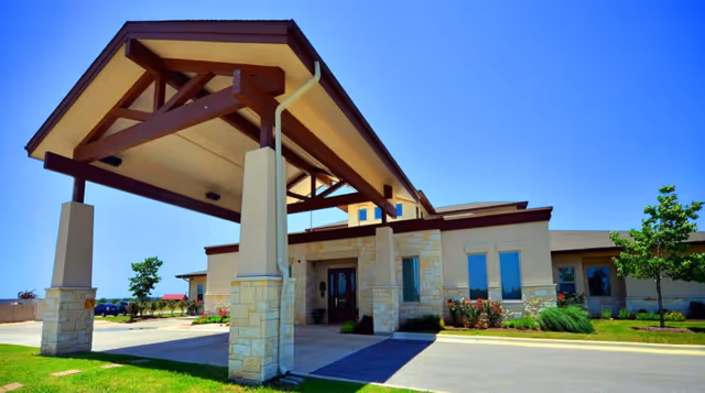 Exterior view of a single-story building with a covered entrance supported by stone pillars and wooden beams, surrounded by a well-maintained lawn and small trees under a clear blue sky.