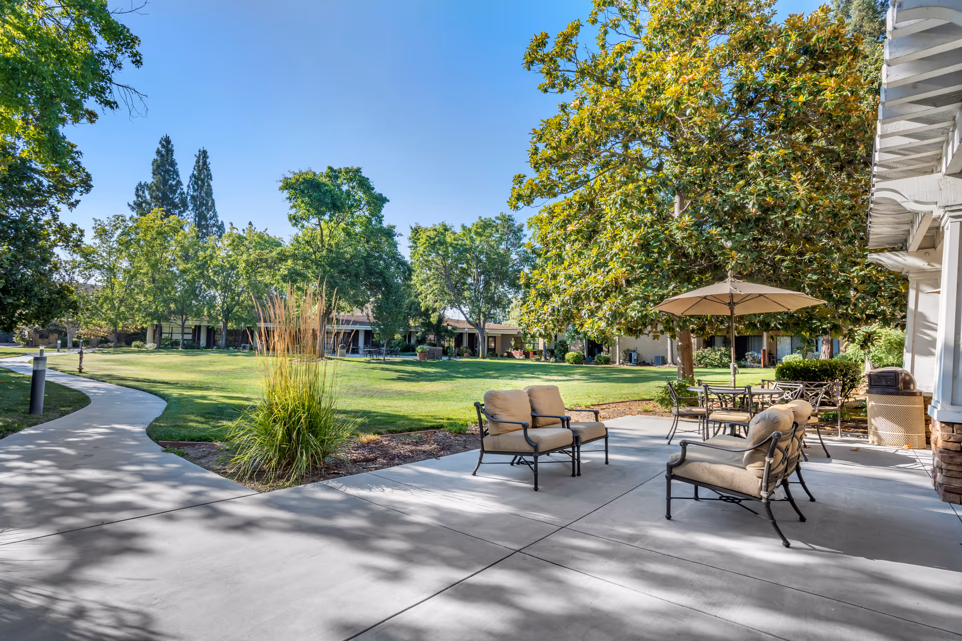 Outdoor patio area with cushioned chairs and a table under a large umbrella, surrounded by green trees and a well-maintained lawn with a curved concrete walkway.