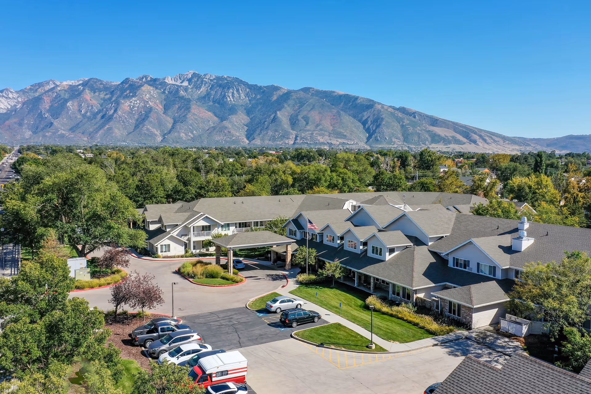 Aerial view of Solstice Senior Living at Sandy facility with a large building surrounded by trees and parking spaces. In the background, there are tall mountains under a clear blue sky.