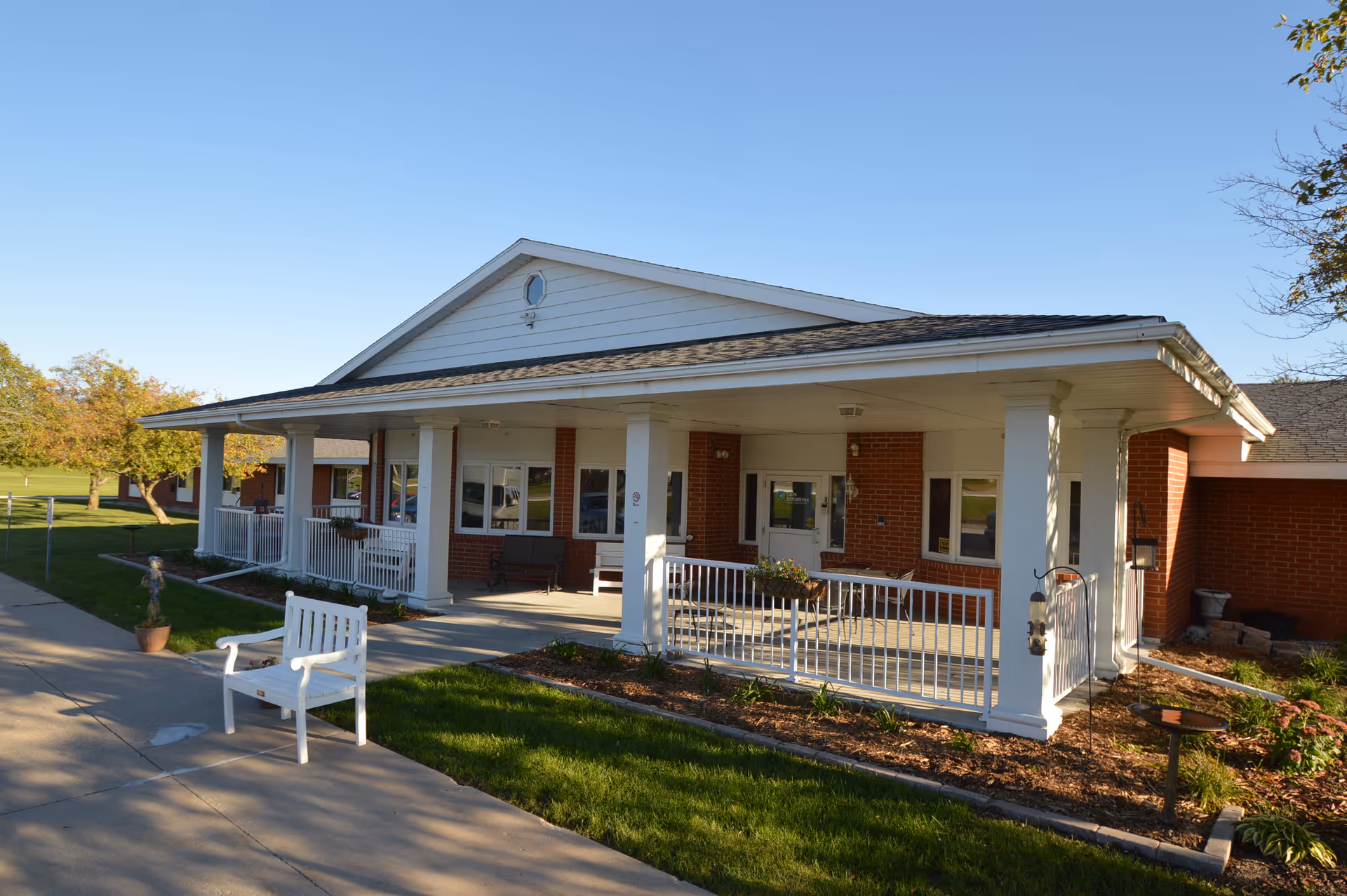 Front exterior view of a single-story brick building with a covered porch supported by white columns. The porch has white railings, benches, and potted plants. The building is surrounded by a well-maintained lawn and trees under a clear blue sky.