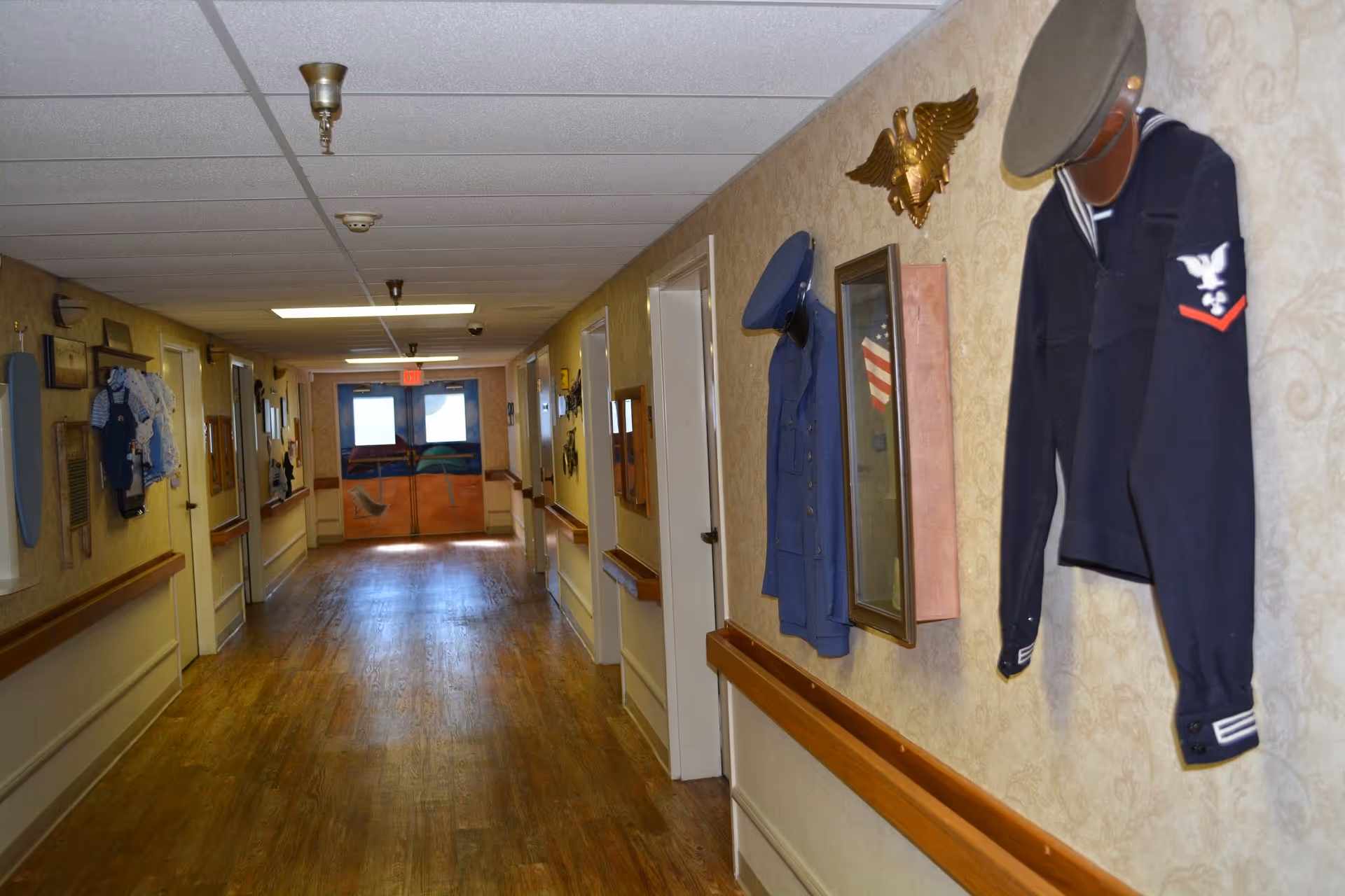 A long hallway with wooden floors and beige walls decorated with military uniforms, hats, and memorabilia. The hallway has handrails on both sides and several doors along the walls. At the end of the hallway, there are double doors with a painted design.