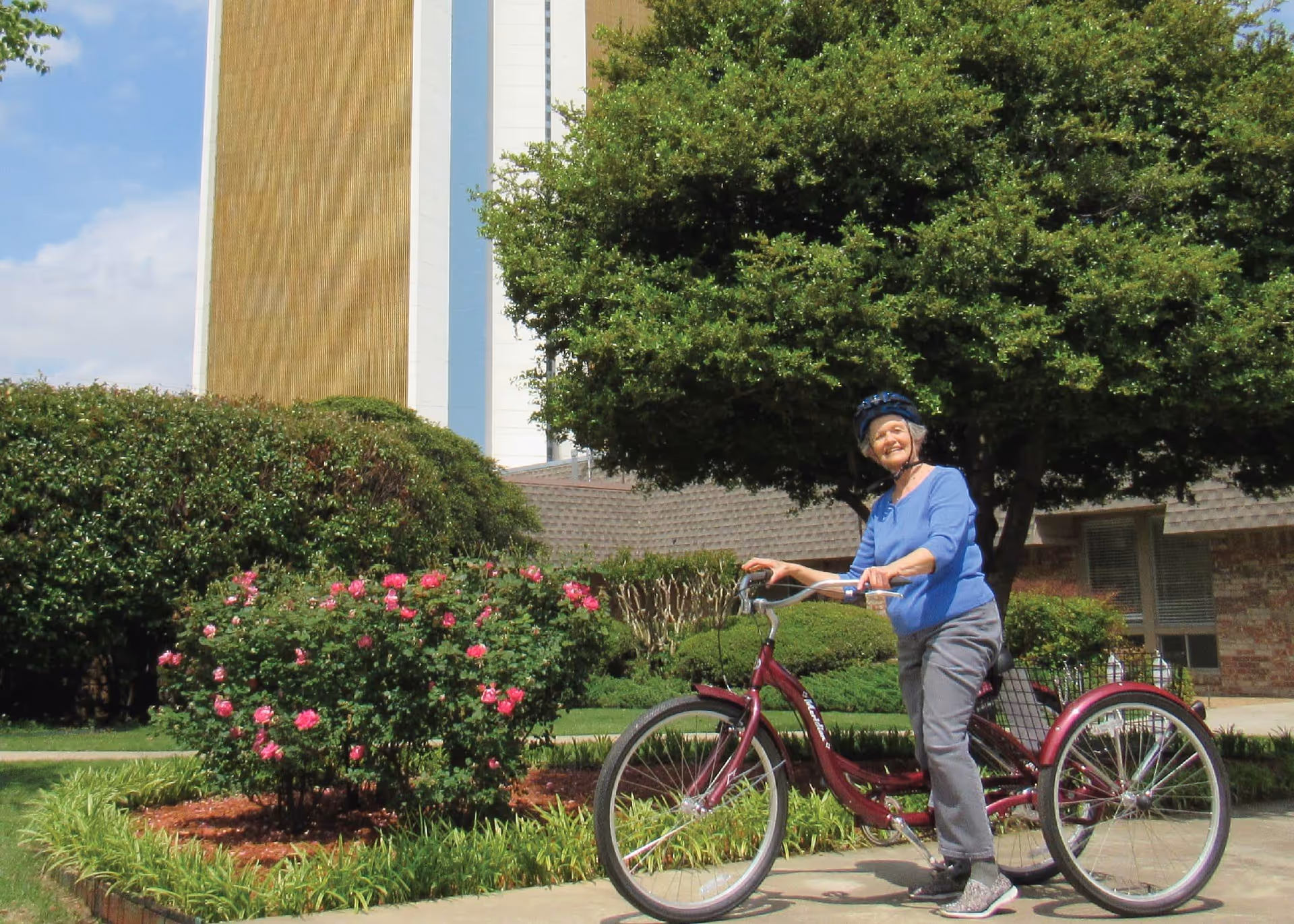 An elderly woman wearing a blue helmet and blue shirt is standing next to a maroon three-wheeled bicycle on a paved path. She is smiling and surrounded by greenery, including bushes with pink flowers and a large tree. In the background, there is a tall building and a smaller brick building.