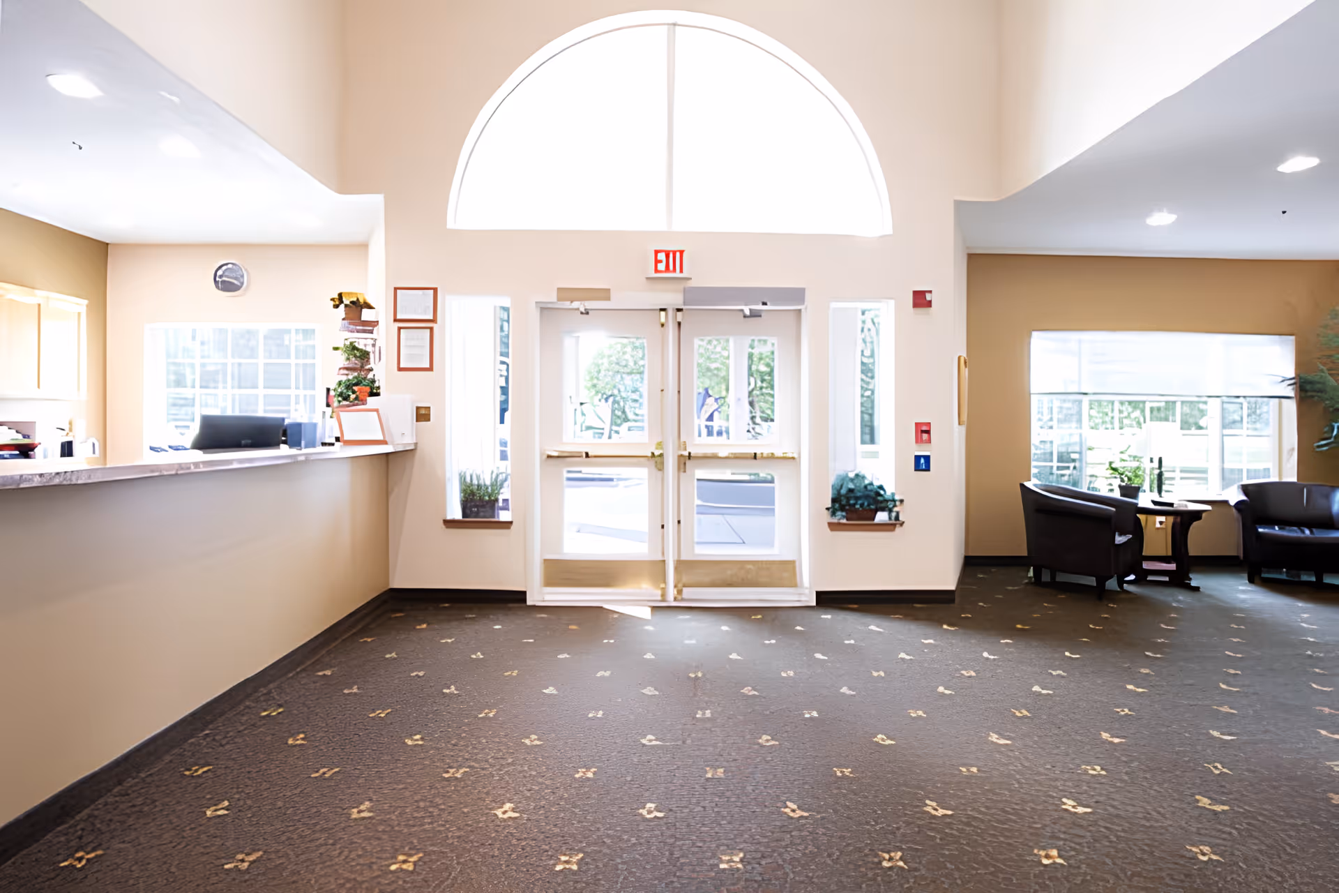 Bright and spacious senior living facility lobby with double glass exit doors under a large arched window. To the left is a reception desk with office equipment and plants, and to the right are seating areas with chairs and a small table near a large window.