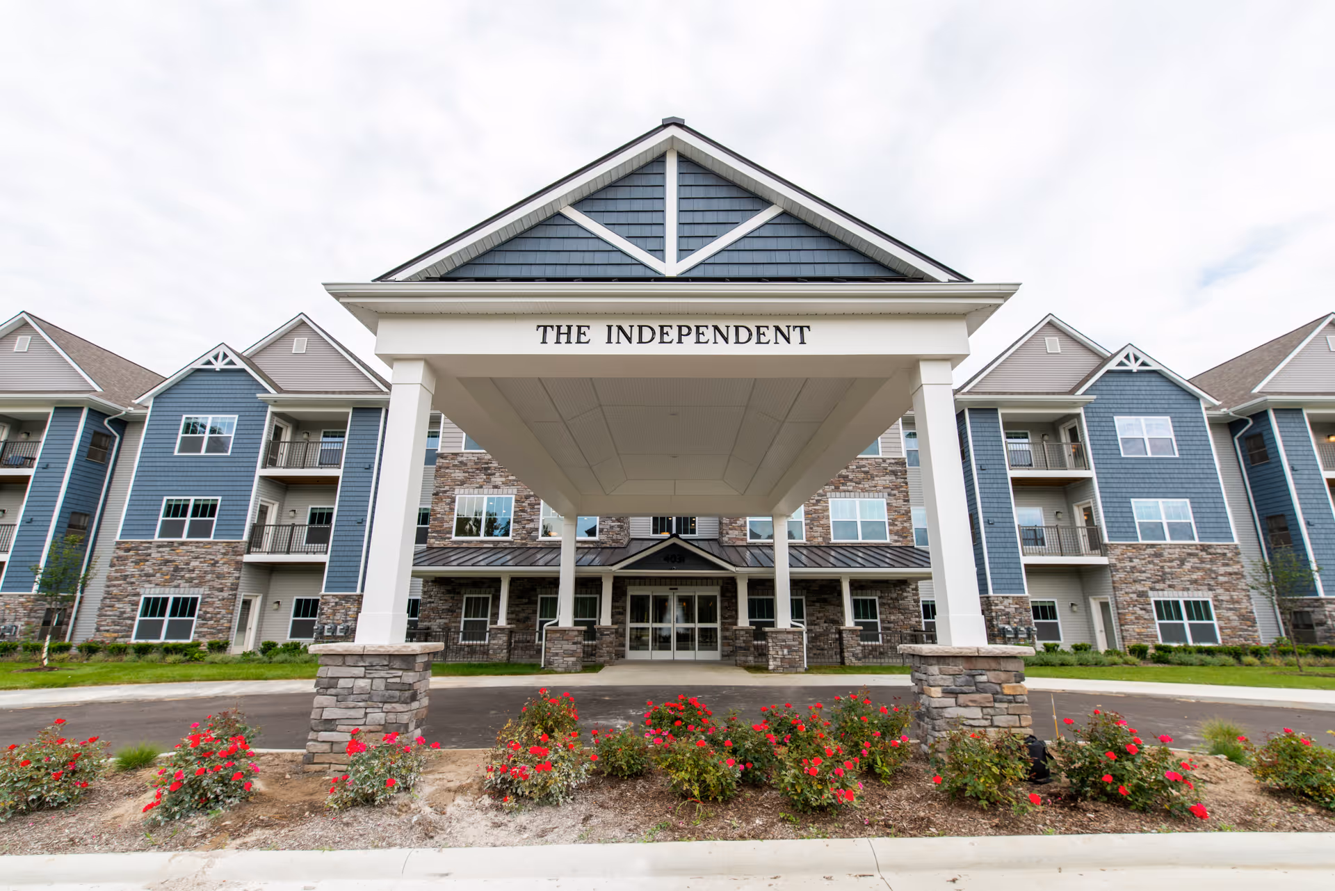 Front entrance canopy of a multi-story retirement building labeled 'THE INDEPENDENT' with stone pillars, balconies, and flowerbeds.