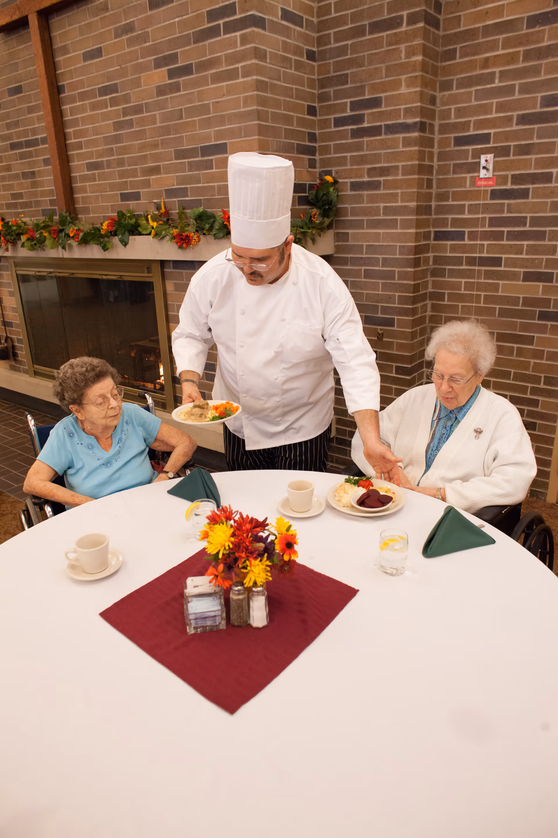 A chef in a white uniform and tall hat serves food to two elderly women seated at a round table with a white tablecloth and a centerpiece of colorful flowers. The setting is indoors with a brick wall and a fireplace decorated with a garland of autumn leaves and flowers in the background.