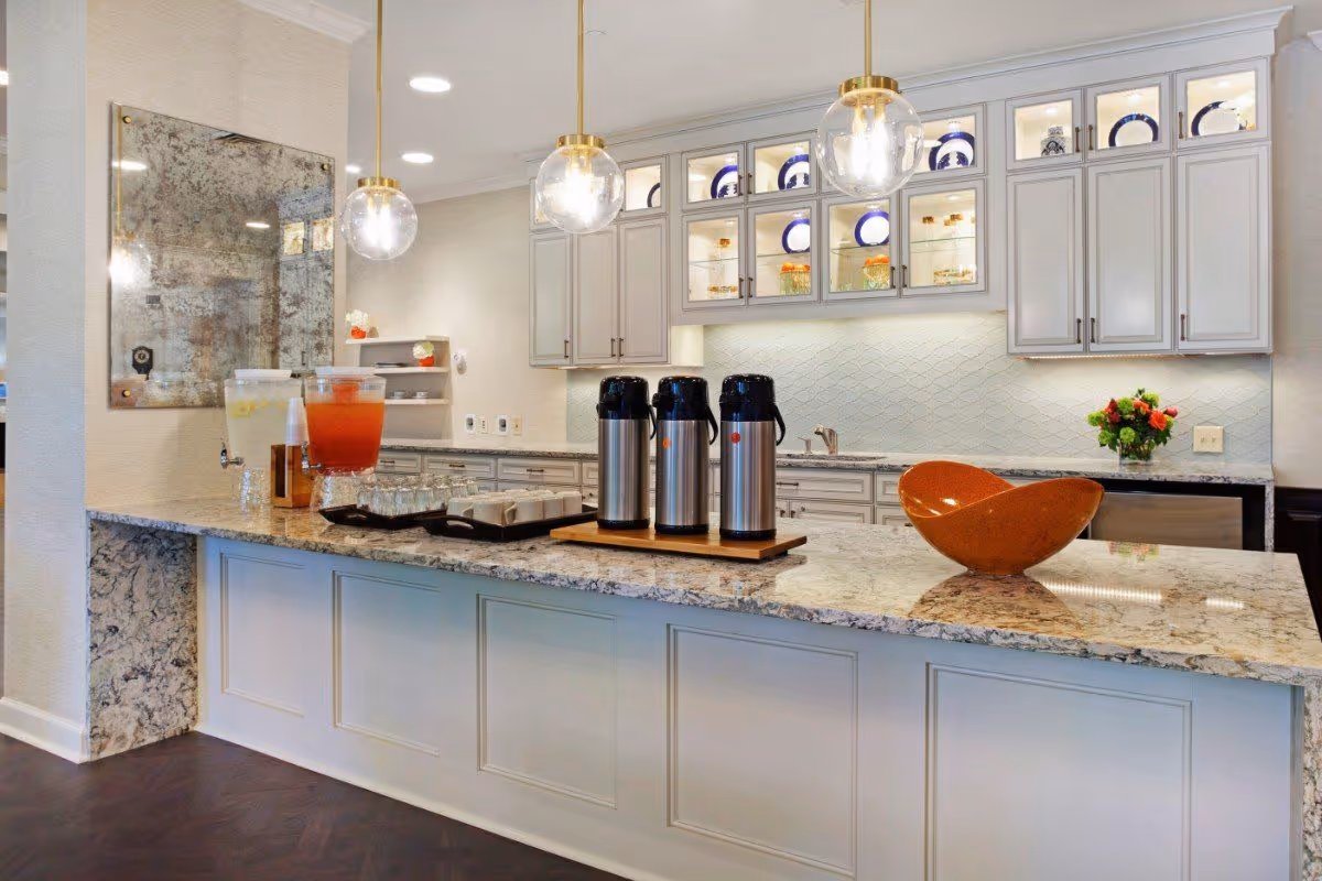 A bright and modern kitchen area with a large granite countertop island. On the island, there are three stainless steel beverage dispensers, a tray with cups, and a large decorative orange bowl. The background features white cabinetry with glass doors displaying blue and white plates, under-cabinet lighting, and a bouquet of flowers on the counter. Two round pendant lights hang from the ceiling above the island.