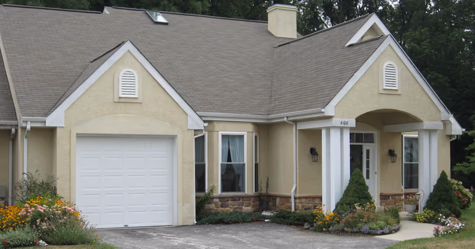 Exterior view of a beige single-story residential building with a gray shingled roof, white garage door, and a covered front entrance supported by white columns. The front yard features neatly trimmed bushes and colorful flowers along the driveway.
