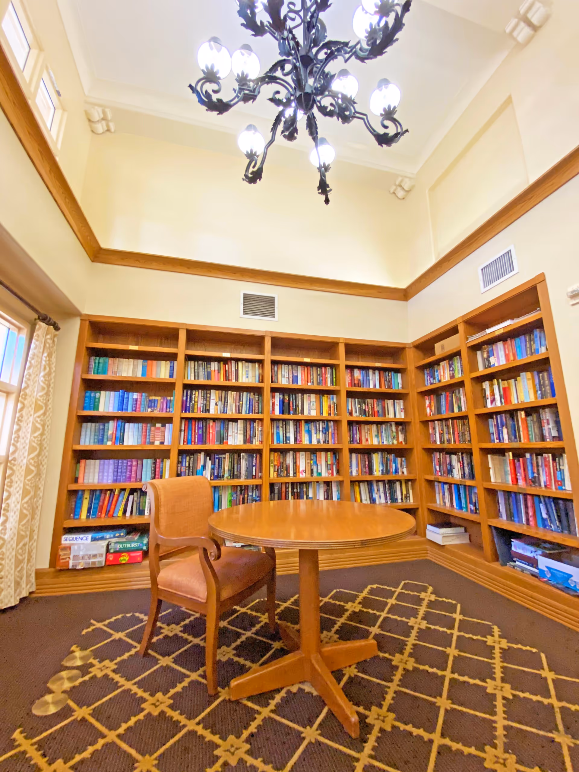 A cozy library room with wooden bookshelves filled with books lining the walls. In the center, there is a round wooden table with a single upholstered wooden chair. The room has a patterned carpet and a decorative chandelier hanging from the high ceiling. A window with patterned curtains is visible on the left side.