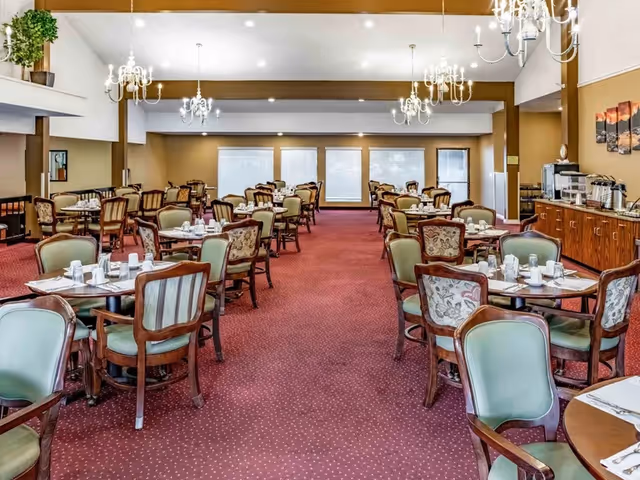 A spacious dining room with multiple round tables set with white cups and napkins. The room features red carpet flooring, green cushioned chairs with wooden frames, chandeliers hanging from the ceiling, and large windows with blinds at the far end. A wooden sideboard with coffee and tea dispensers is visible on the right side.
