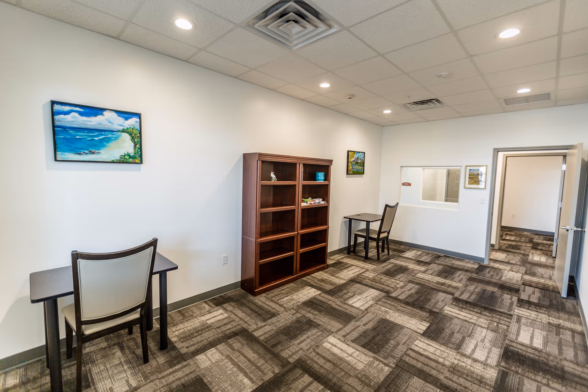 A bright interior room with two small desks and chairs placed against white walls. There is a wooden bookshelf with some items on it between the desks. The room has patterned carpet flooring and recessed ceiling lights. Three framed paintings hang on the walls, and an open doorway leads to another room.