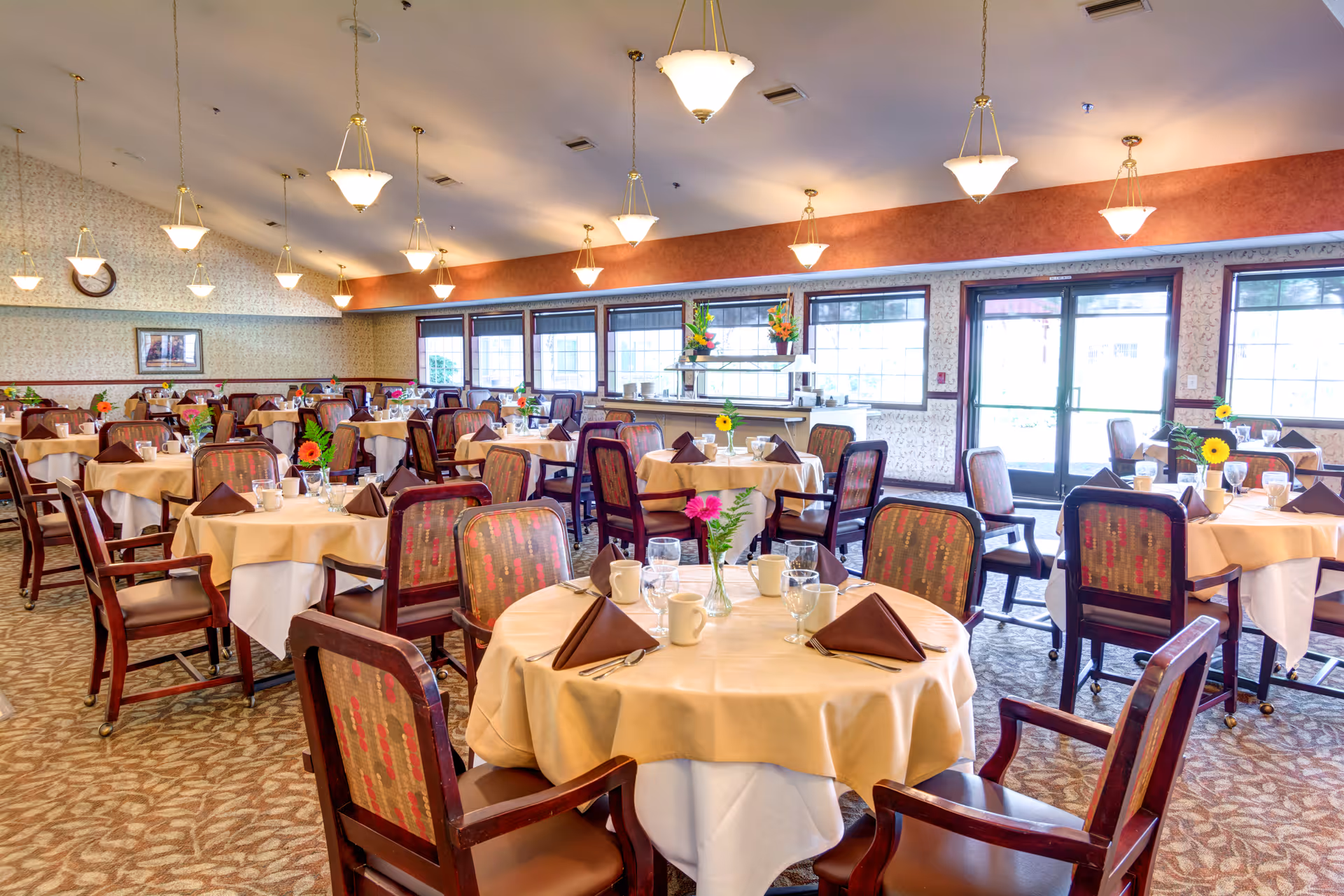 Spacious retirement community dining room with round tables set with tablecloths, napkins, glassware, and flower centerpieces.