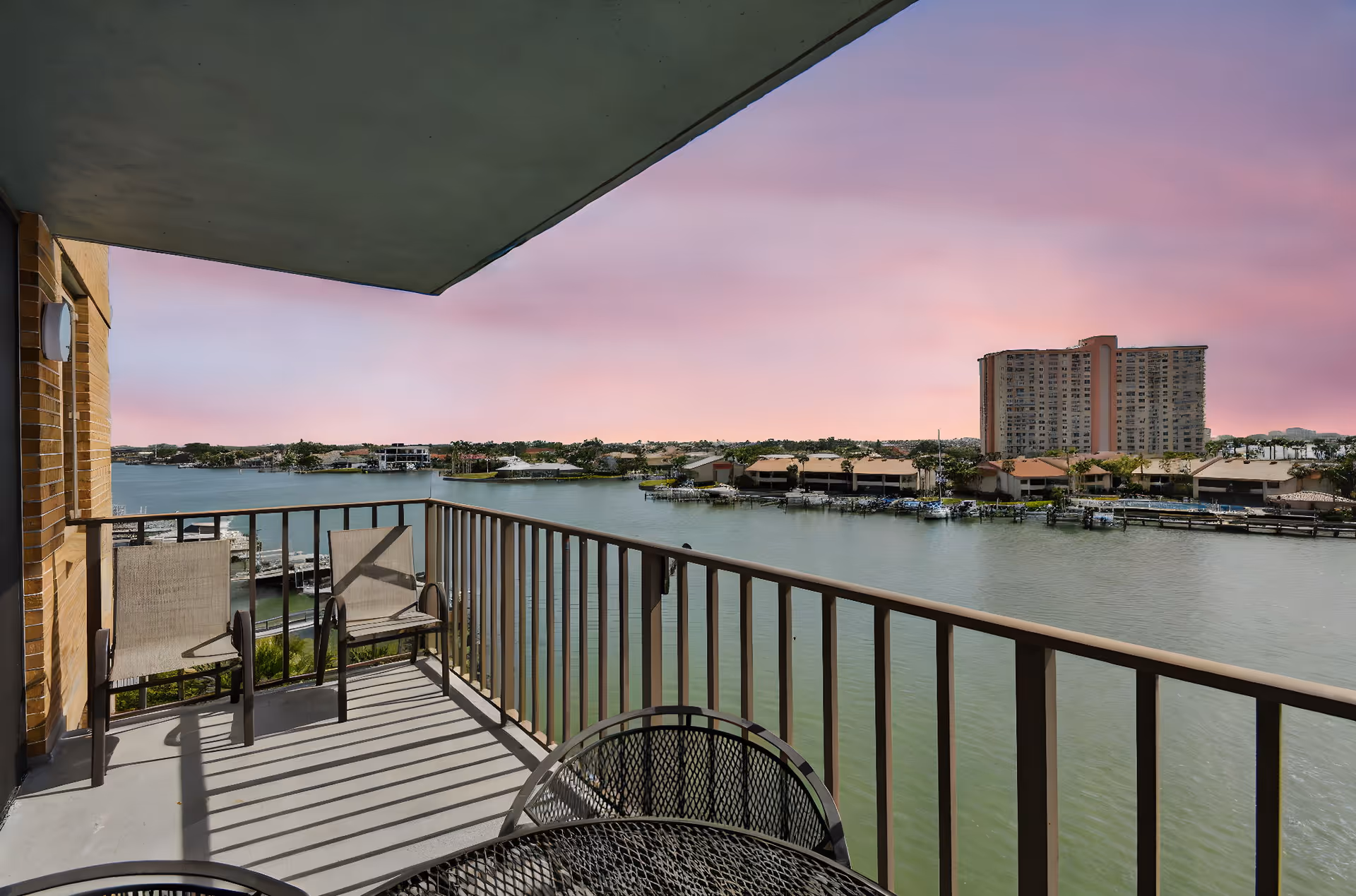 View from a balcony overlooking a calm body of water with a railing, two chairs, and a round metal table. Across the water, there are houses with docks and a tall apartment building under a pink and purple sky.