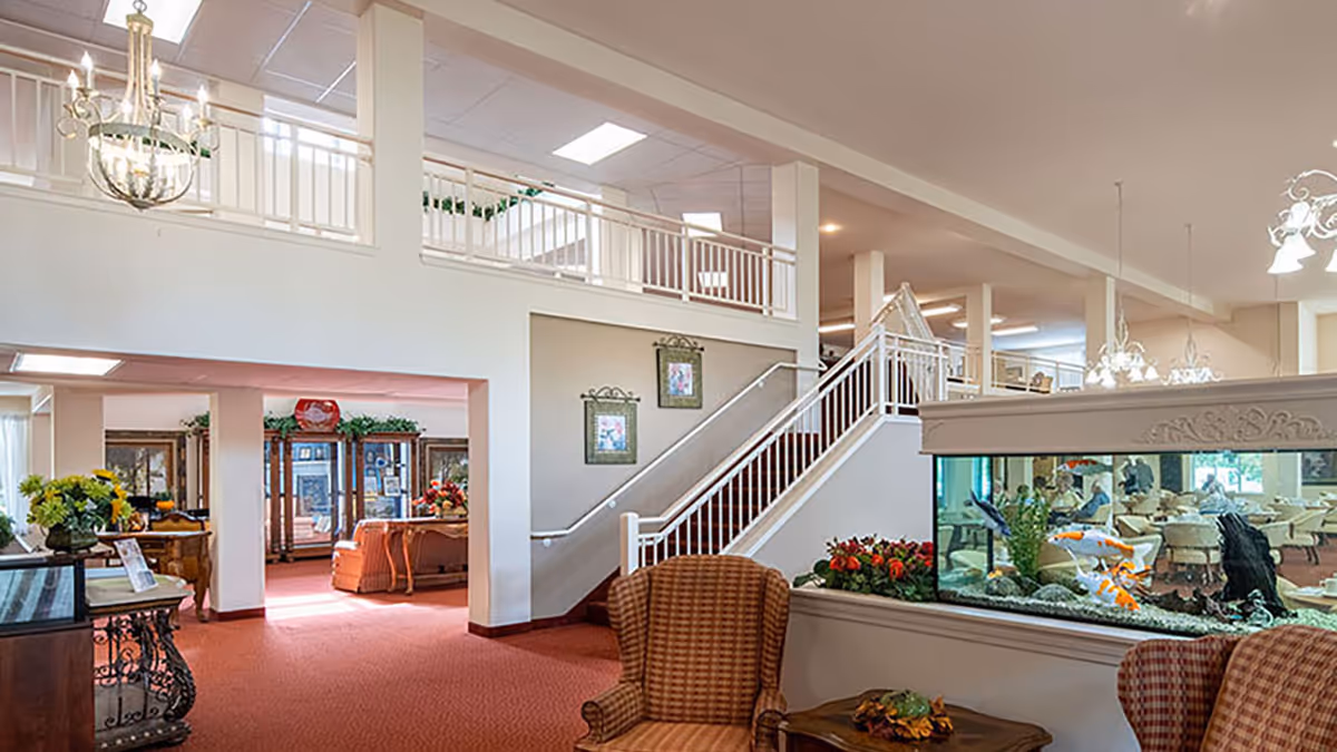 Interior view of a senior living facility lobby area with a staircase leading to an upper floor. The space features comfortable seating with patterned armchairs, a large fish tank with colorful koi fish, decorative plants, and framed artwork on the walls. The area is well-lit with chandeliers and ceiling lights, and a dining area is visible in the background.