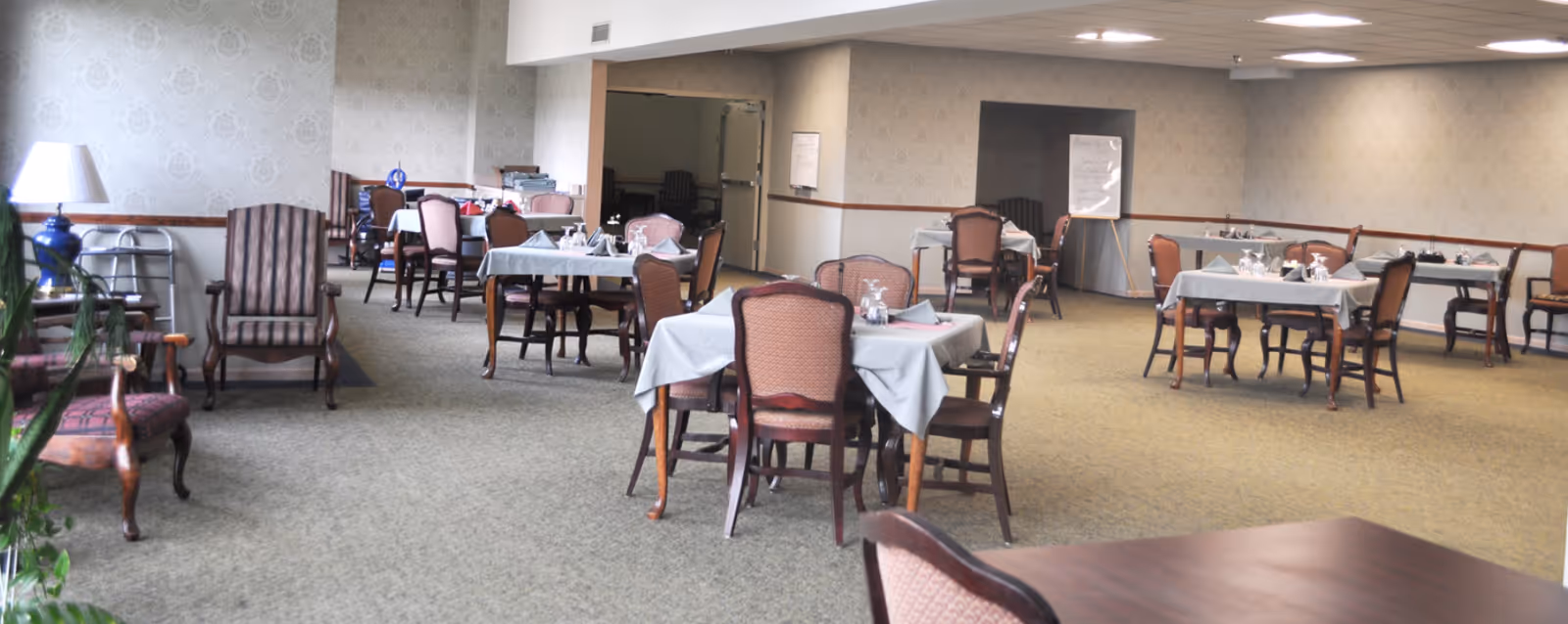 Spacious dining room with multiple set tables, upholstered chairs, and a few lounge chairs in a senior care facility.