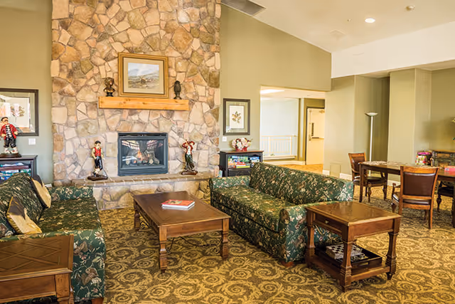 A cozy living room area with two green floral patterned sofas facing each other across a wooden coffee table. The room features a stone fireplace with decorative figurines and a framed painting above the mantel. There are side tables next to the sofas and a dining table with chairs in the background. The carpet has a patterned design and the walls are painted in neutral tones.