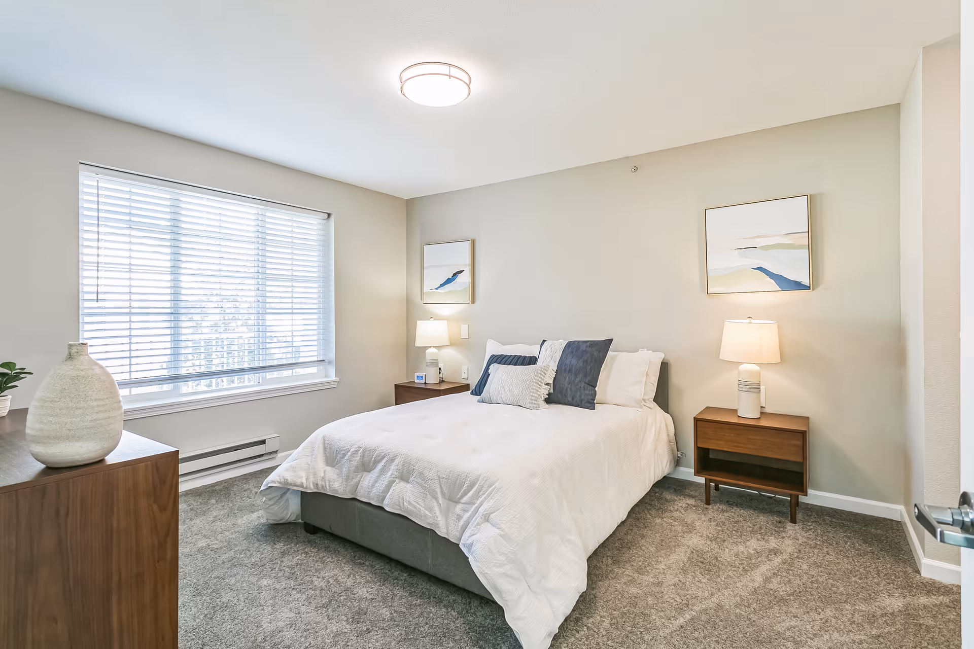 Bright bedroom with a neatly made bed, two nightstands with lamps, artwork on the walls, and a large window with blinds.