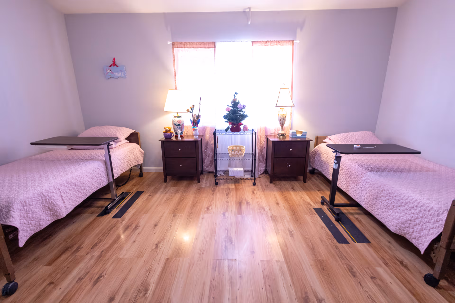 A bright room with two single beds on either side, each covered with a pink quilt. Each bed has an adjustable overbed table. Between the beds are two dark wooden nightstands with lamps and decorative items. A small metal shelving unit with a small Christmas tree and basket is placed in front of a window with sheer pink curtains. The floor is light wood.
