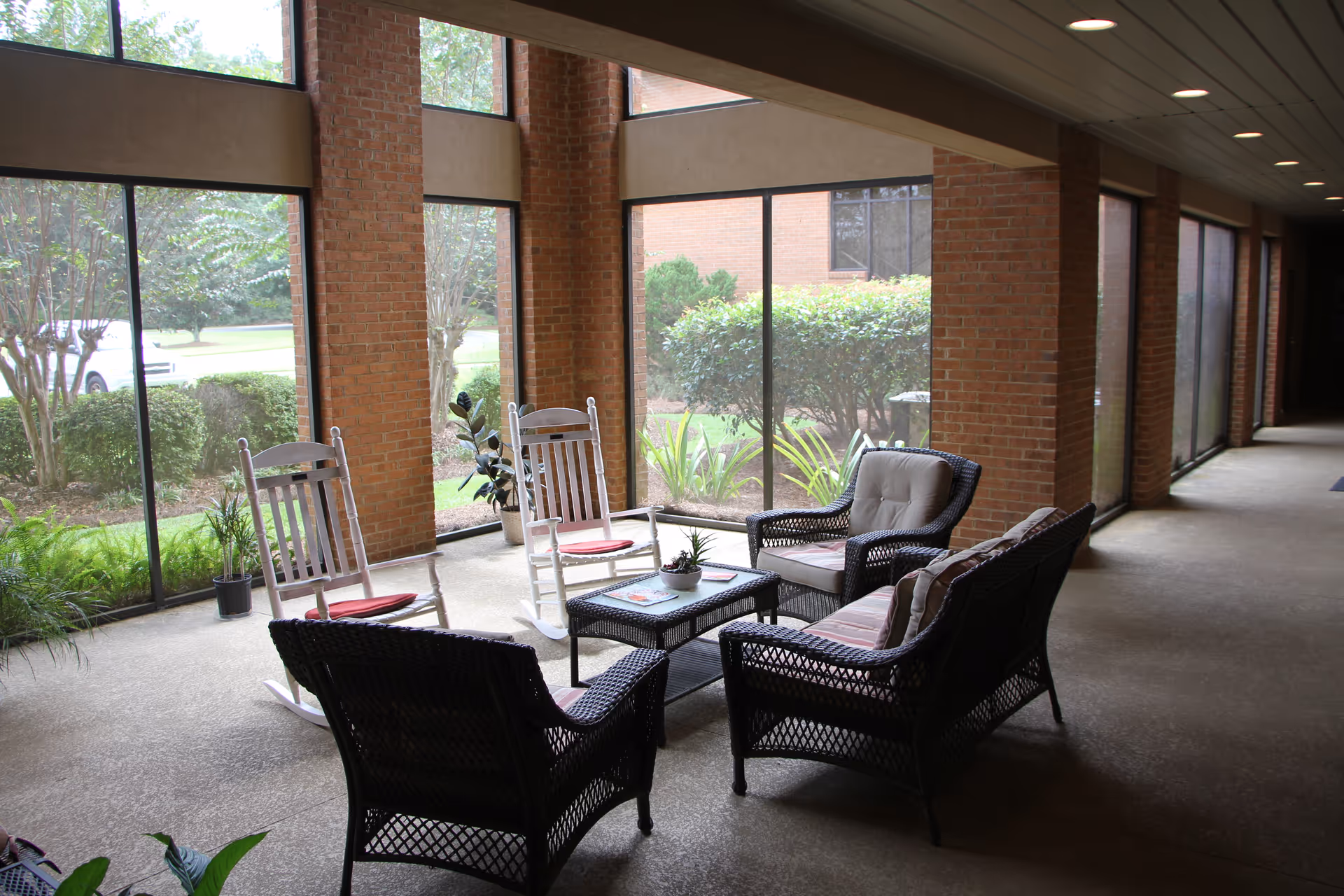 A bright indoor seating area with large floor-to-ceiling windows showing greenery outside. The space features two white rocking chairs with red cushions, two black wicker chairs with cushions, and a black wicker loveseat with cushions arranged around a small black coffee table with a plant and magazines on it. The walls are made of red brick and the floor is a light-colored textured surface.