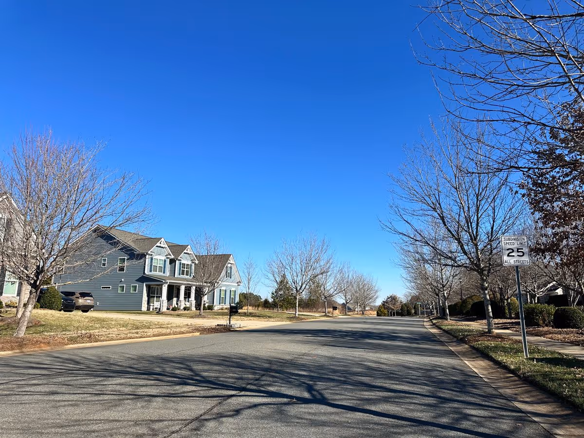 A quiet suburban street lined with leafless trees and two-story houses under a clear blue sky.