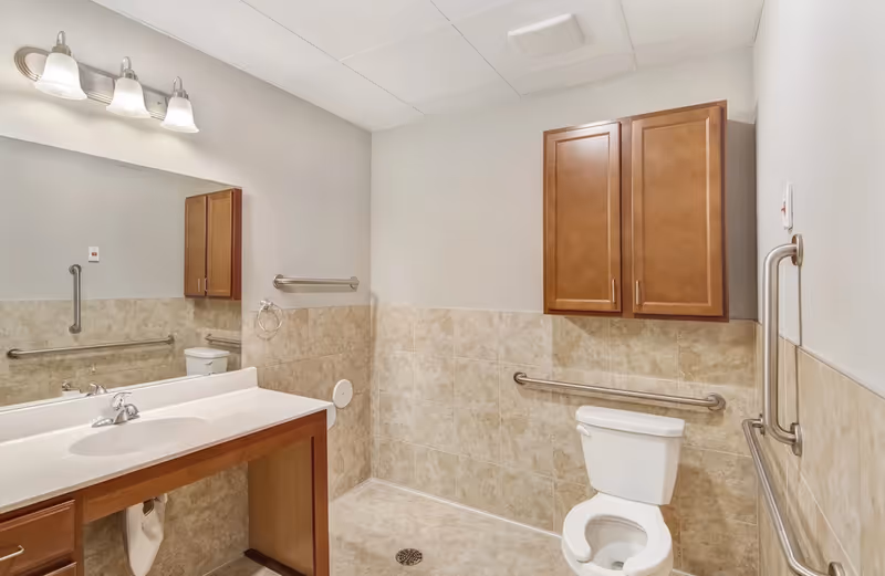 A clean and accessible bathroom with beige tiled walls and floor. The bathroom features a white toilet with grab bars on the walls for support, a wooden cabinet mounted above the toilet, a large mirror above a white sink with a wooden vanity, and three light fixtures above the mirror.