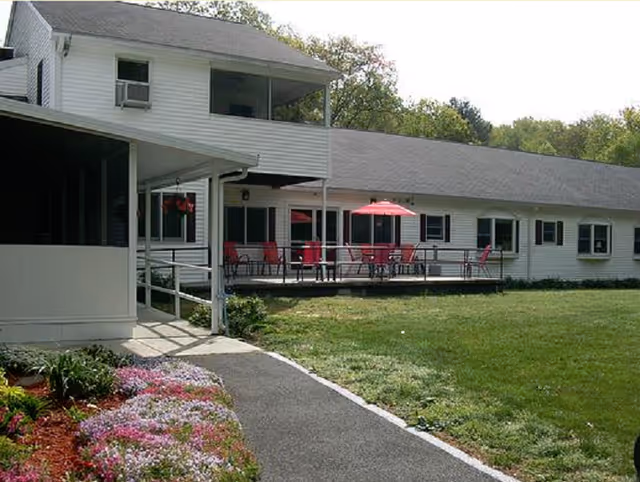 Exterior view of a single-story white building with a gray roof, featuring a patio area with red chairs and a red umbrella. There is a paved walkway leading to the entrance, surrounded by a garden with colorful flowers and a grassy lawn. Trees are visible in the background.