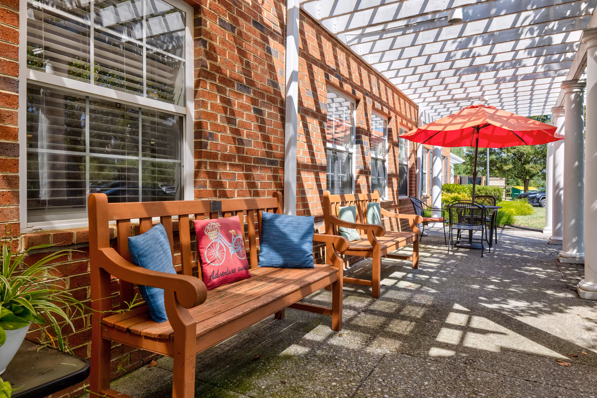 Outdoor patio area with wooden benches adorned with colorful cushions, a table with chairs under a red umbrella, and a brick wall with windows. The area is shaded by a white pergola casting shadows on the ground.
