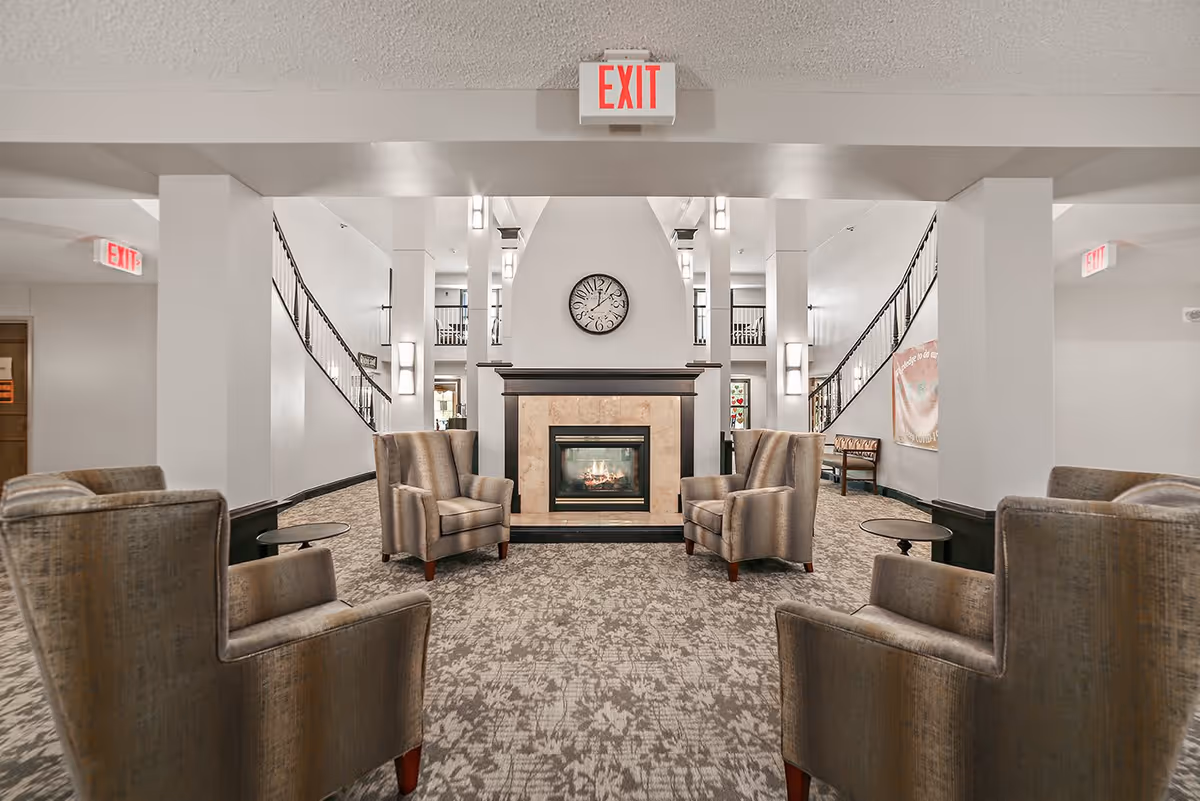 A spacious and well-lit common area with a central fireplace and a clock above it. Four upholstered armchairs are arranged around the fireplace, with small round tables beside some of them. The room features carpeted flooring, white walls, and two staircases with black railings on either side leading to an upper level. Multiple exit signs are visible on the ceiling and walls.