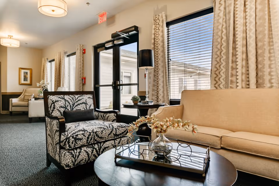 Comfortable senior living lounge with a patterned armchair, beige sofa, coffee table with a vase, large windows, and a glass exit door.