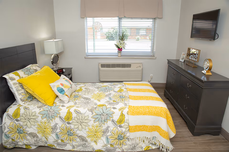 Well-lit bedroom with a floral bedspread, yellow pillows and striped throw, a dresser with decor and a wall-mounted TV, and a window above an air conditioning unit.