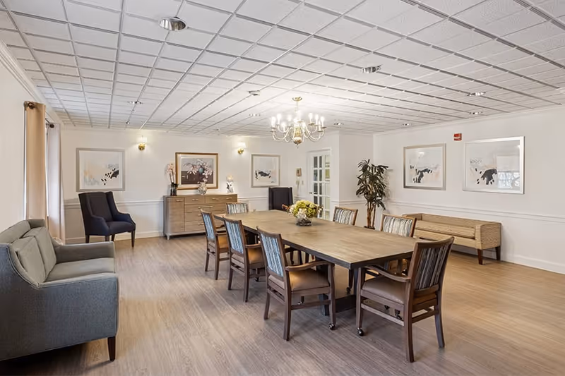 A spacious, well-lit room with a long wooden table surrounded by eight chairs. The room features light-colored walls adorned with framed artwork, a chandelier hanging from the ceiling, a gray sofa on the left, two dark armchairs, a wooden sideboard with decorative items, a potted plant, and a bench along the right wall. The floor is light wood, and the ceiling has a grid pattern with recessed lighting.