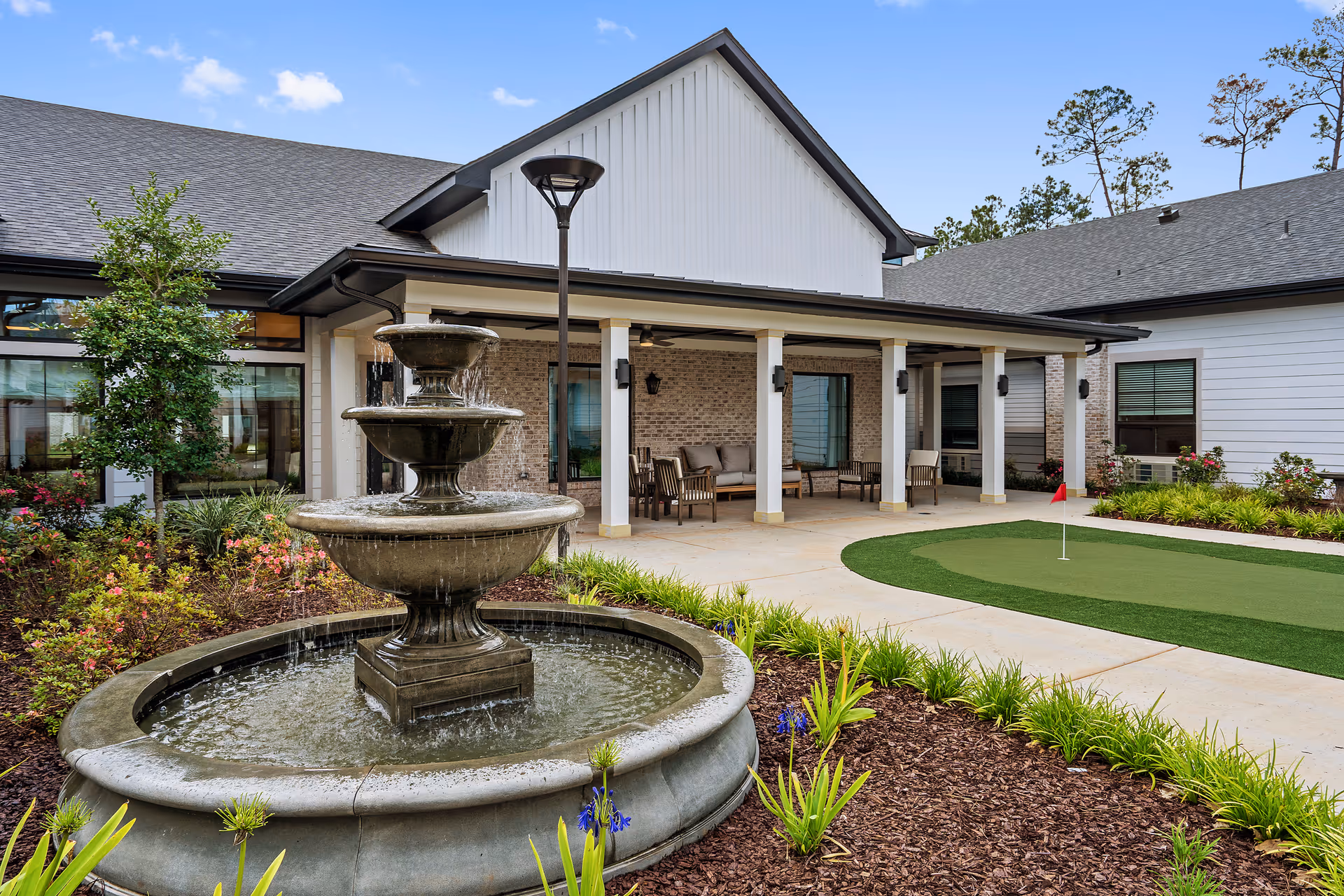 Outdoor courtyard area at Sage Lake featuring a three-tiered water fountain surrounded by landscaped plants and mulch, a putting green with a red flag, and a covered patio with seating including chairs and a sofa. The building has white siding, brick accents, and a dark roof under a blue sky with a few clouds.