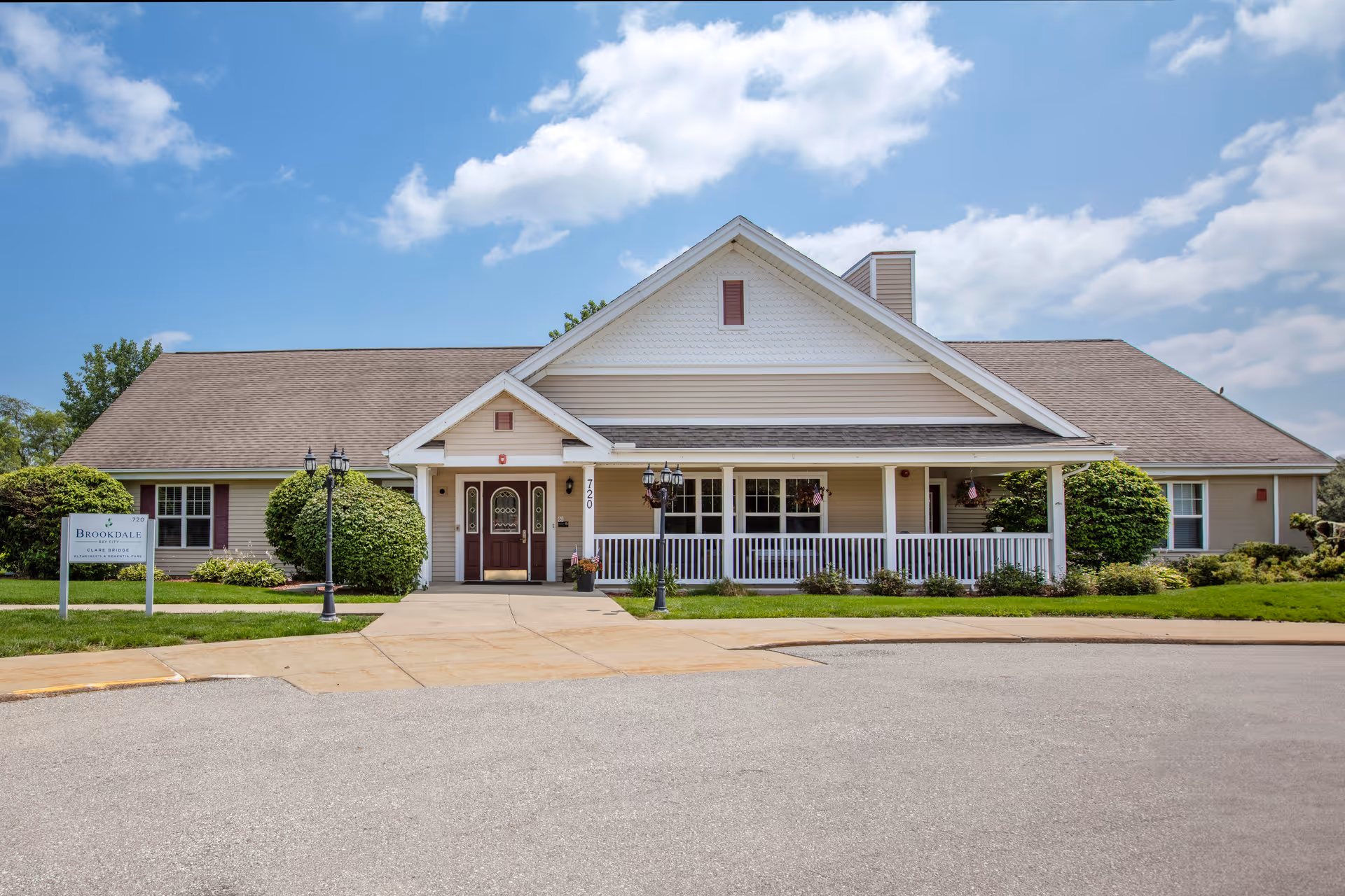 Front exterior view of a single-story building with beige siding and a brown roof under a partly cloudy blue sky. The building has a covered porch with white railings, a double-door entrance, and well-maintained landscaping with bushes and grass. A sign on the left side reads 'Brookdale'.