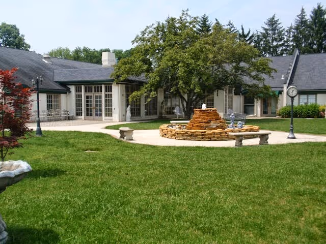 Outdoor courtyard area of a senior living facility with a circular stone fountain in the center, surrounded by benches and a large tree. The building with multiple windows and doors is visible in the background, along with green grass and a few lampposts.