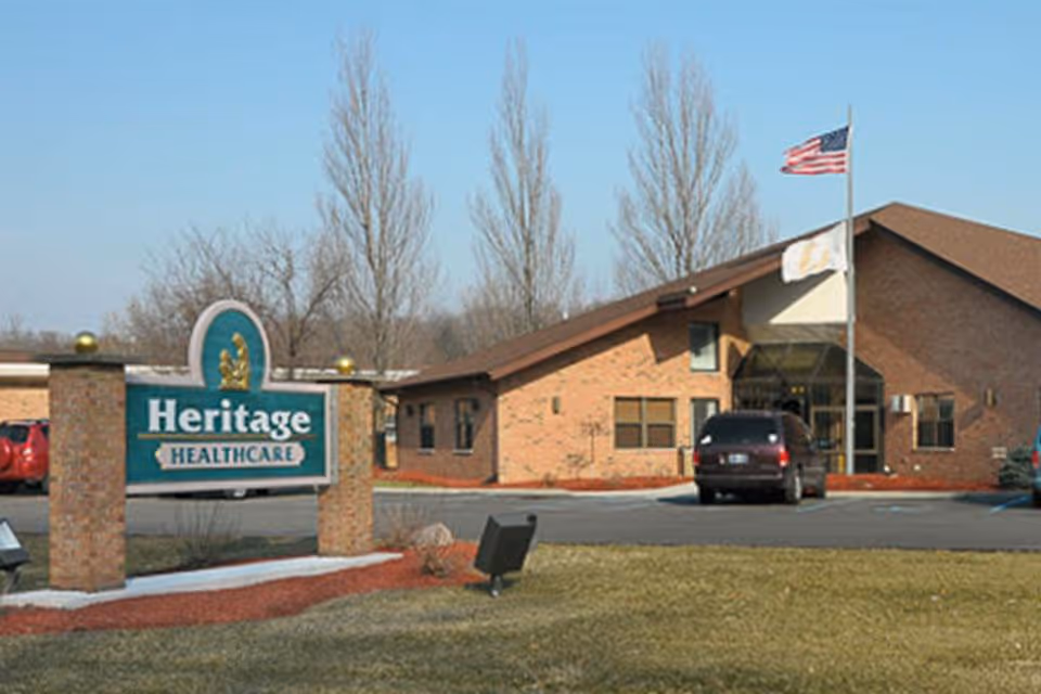 Exterior view of Heritage Healthcare facility showing a brick building with a sloped roof, an American flag and another flag on flagpoles, a parking area with vehicles, and a sign in front that reads 'Heritage Healthcare'.