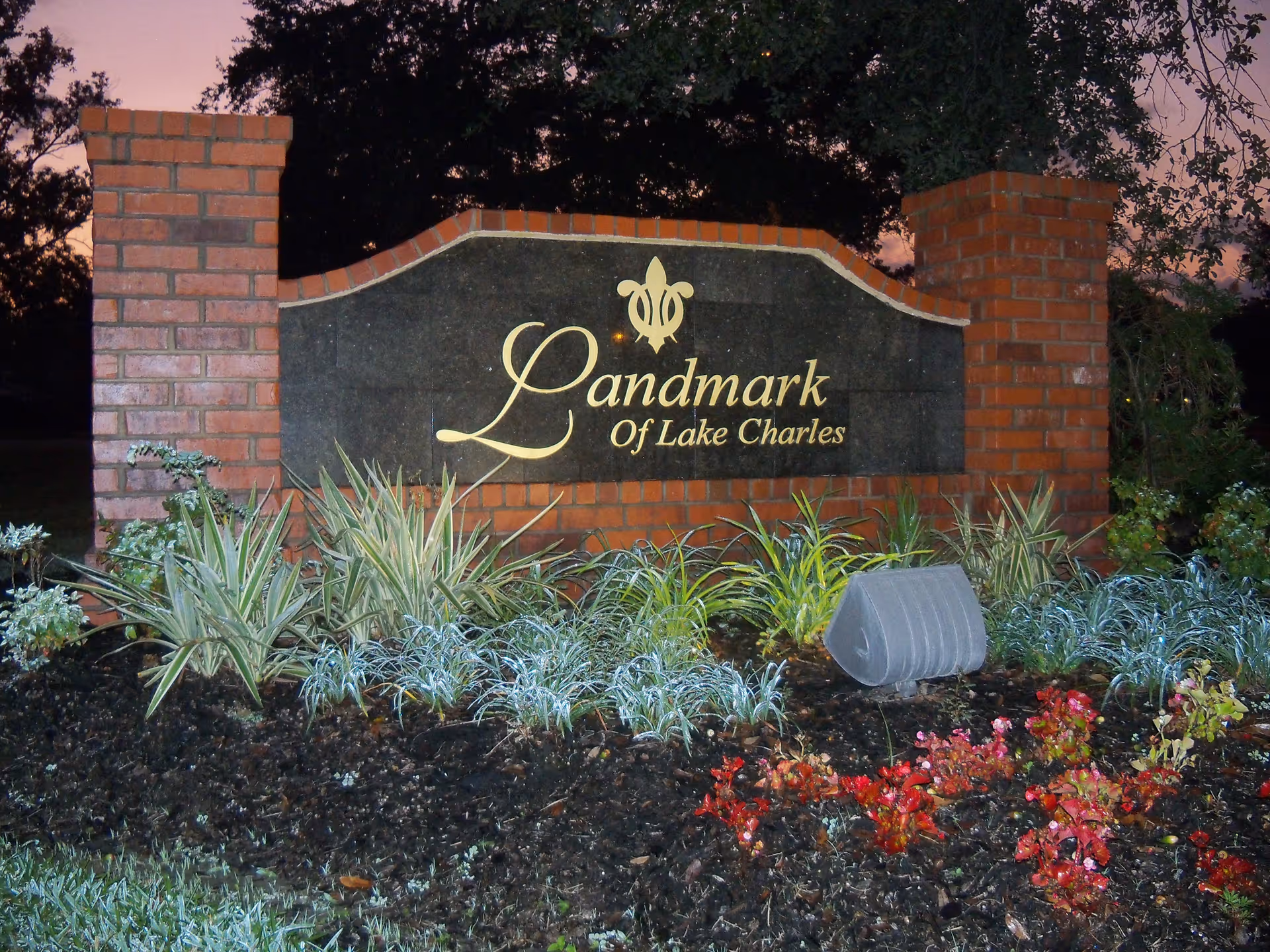 A brick and stone sign for Landmark of Lake Charles surrounded by plants and flowers, with trees and a dusky sky in the background.