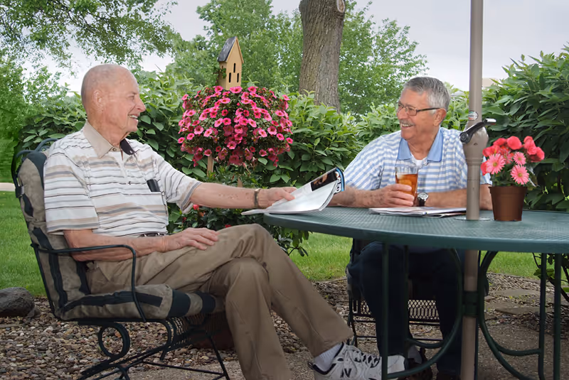 Two elderly men sitting outdoors at a round green metal table with an umbrella pole in the center. One man is holding a newspaper and the other is holding a glass of iced tea. They are smiling and engaged in conversation. Behind them are green bushes and a tree, with a hanging basket of pink flowers and a small birdhouse.