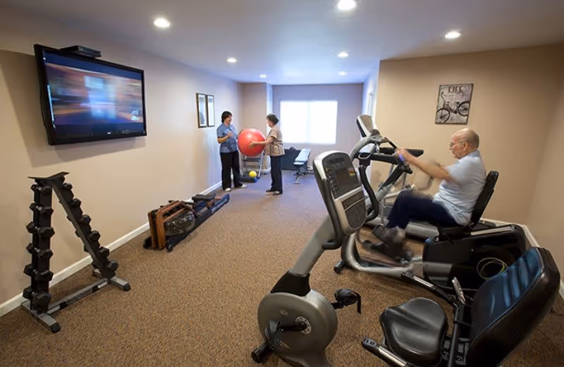 A fitness room in a senior living facility with exercise equipment including stationary bikes and weights. An elderly man is using a recumbent bike while two women stand near a large red exercise ball in the background. A flat-screen TV is mounted on the wall, and the room is carpeted with beige walls and ceiling lights.