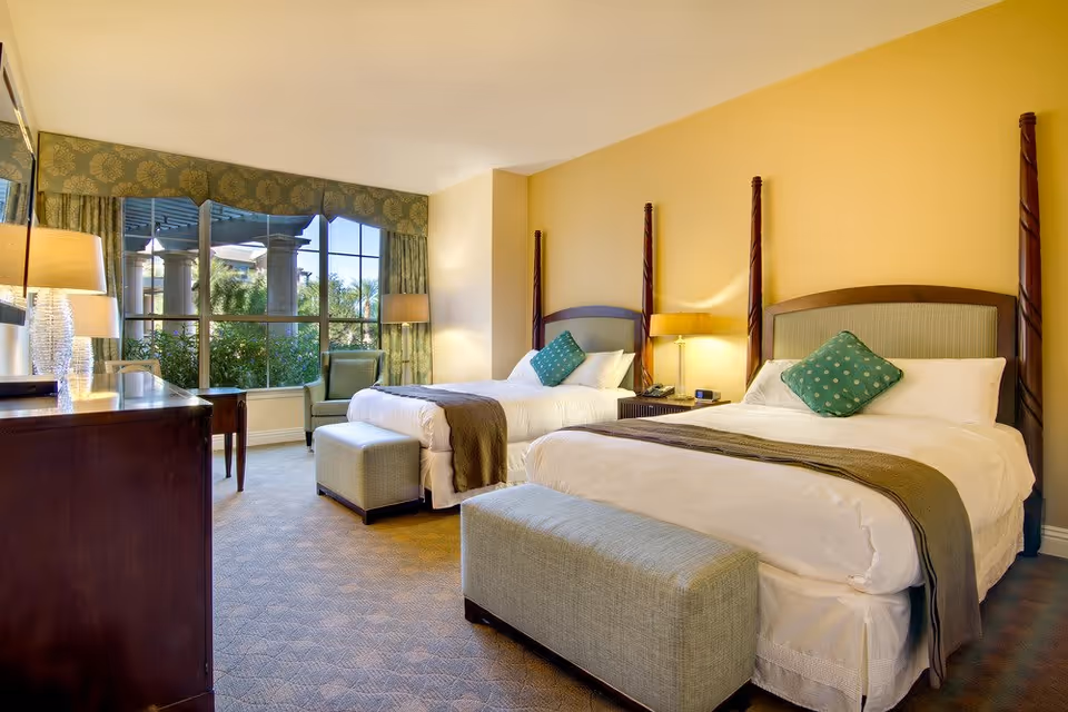 A bright and spacious bedroom with two twin beds, each with a green decorative pillow and a gray throw blanket. At the foot of each bed is a cushioned bench. The room features a large window with green patterned curtains, a green armchair, two bedside tables with lamps, and a wooden dresser with a lamp on top.
