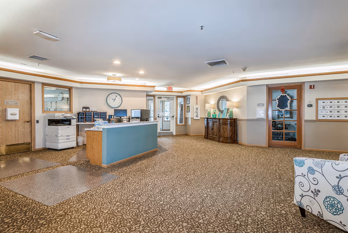 Reception area of a senior living facility with a front desk, office equipment, a clock on the wall, a wooden cabinet with lamps and decorative items, patterned carpet, and a floral upholstered chair partially visible.