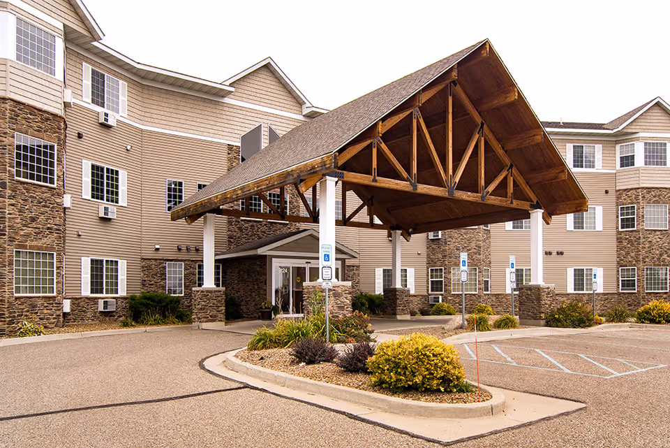 Exterior view of a senior living facility with a large covered entrance supported by white columns and stone bases. The building has multiple windows and a combination of stone and beige siding. There are several handicap parking spaces in front of the entrance.