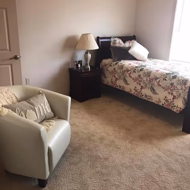 Bedroom with a floral twin bed, wooden nightstand and lamp, and a beige armchair with pillows.
