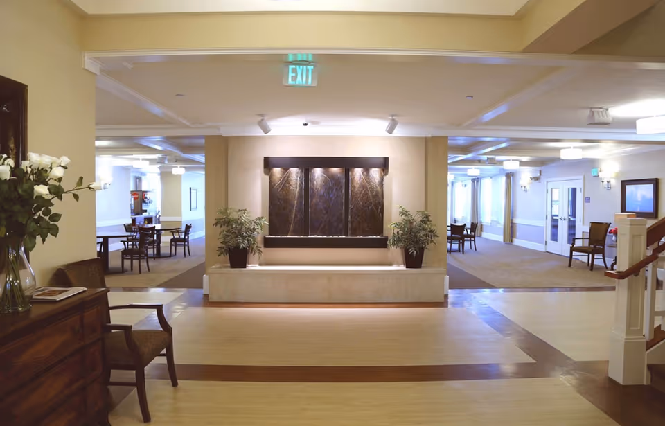 Interior view of a senior living facility common area with a water feature on the far wall flanked by two potted plants. There are chairs and tables arranged in the background on both sides, soft lighting, and an exit sign above the water feature. A wooden chest with a vase of white flowers and a chair are visible on the left side, and a staircase railing is visible on the right.