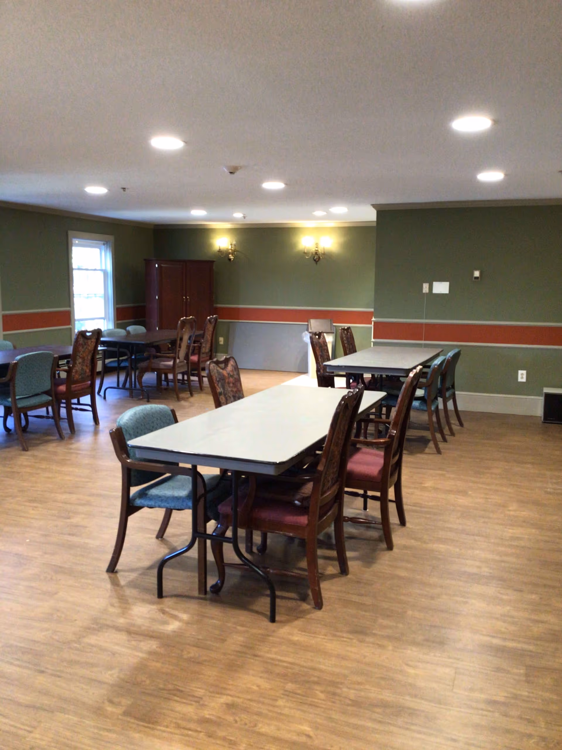 A dining room with several rectangular tables and a mix of wooden chairs with different upholstery patterns. The walls are painted green with a red and beige horizontal stripe. There are two wall sconces with lights on the far wall and a window on the left side letting in natural light. The floor is wood, and the ceiling has multiple recessed lights.
