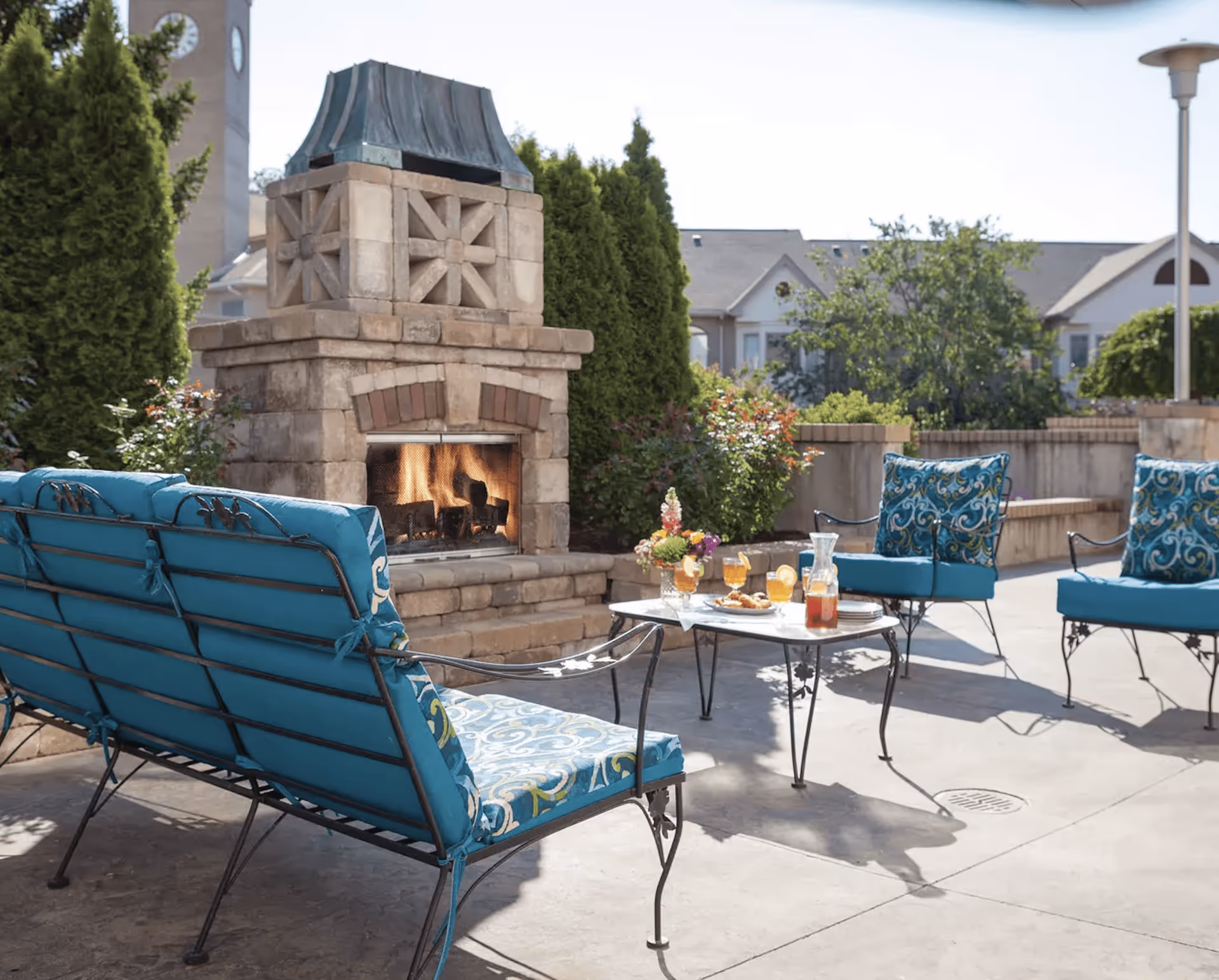 Outdoor patio area with a stone fireplace, surrounded by blue cushioned metal chairs and a loveseat. A small table in the center holds a pitcher of iced tea, glasses with lemon slices, and a plate of snacks. Green shrubs and trees are visible in the background along with residential buildings.