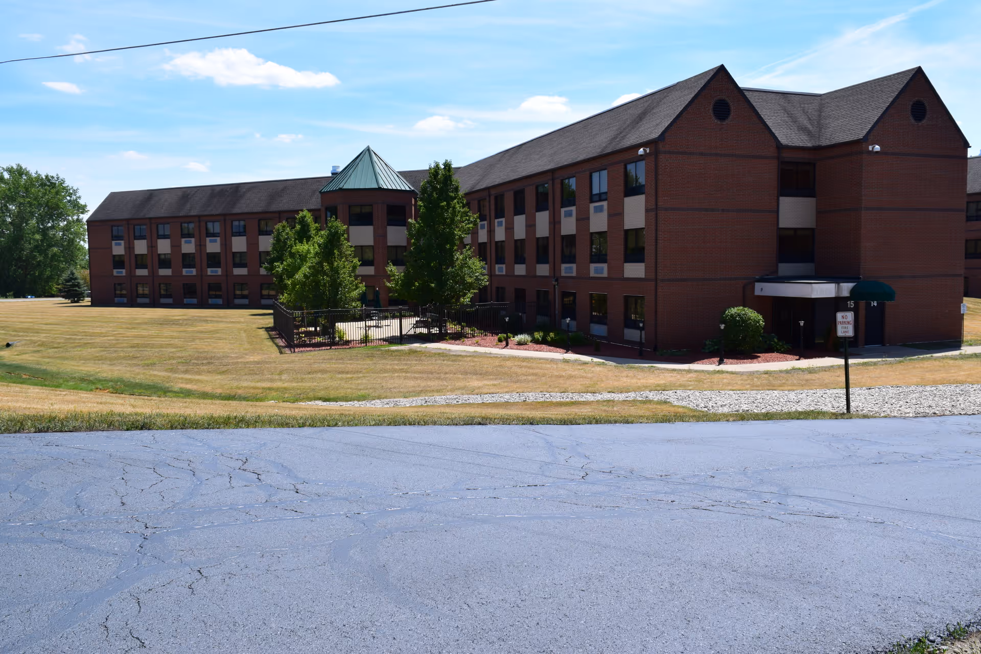 Exterior view of a three-story brick building with multiple windows, surrounded by a grassy area and a paved driveway in the foreground. The building has a small fenced garden area with trees and a covered entrance.
