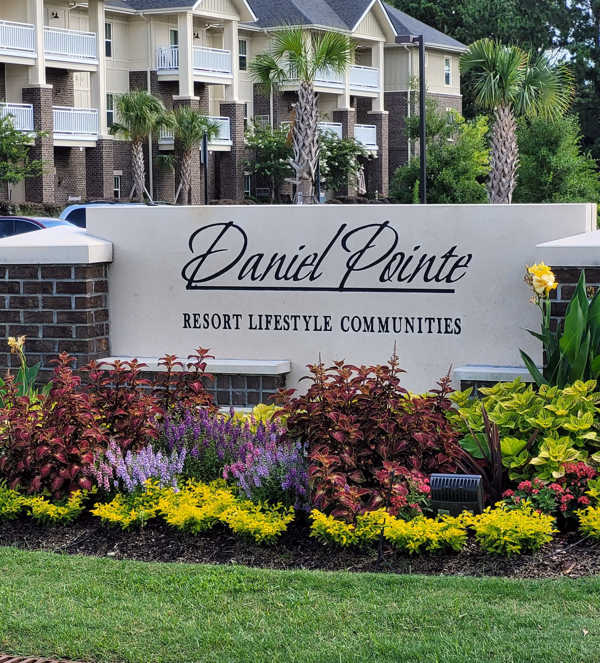 Entrance sign for Daniel Pointe Resort Lifestyle Communities surrounded by colorful flowers and greenery, with a multi-story residential building and palm trees in the background.