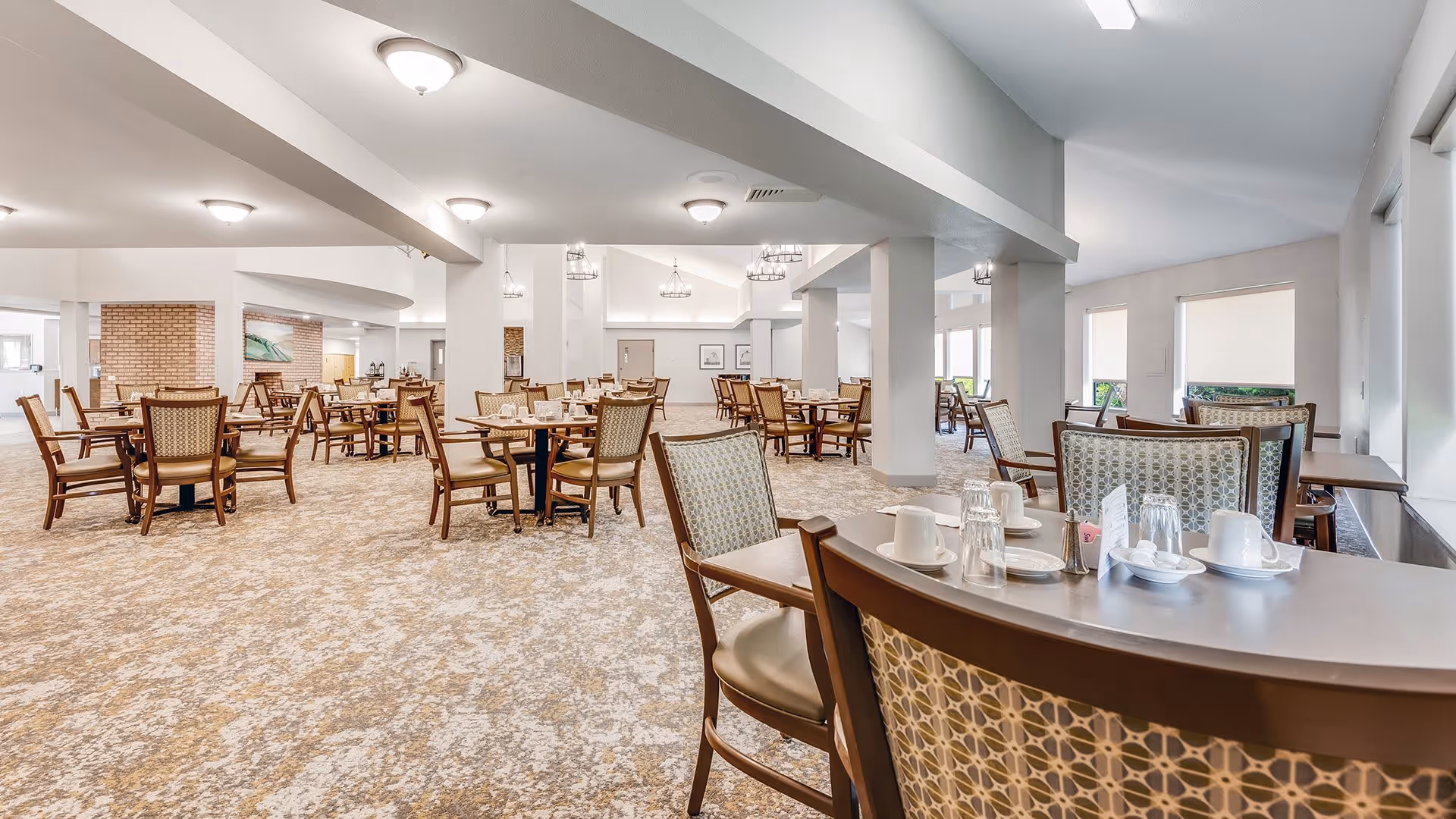 A spacious dining room with multiple wooden tables and chairs arranged neatly. The tables are set with white cups, plates, and glasses. The room has a beige patterned carpet, white walls, and several ceiling lights providing bright illumination. Large windows with blinds allow natural light to enter the space.