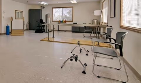 A spacious room with exercise equipment including parallel bars, chairs, and a walker, likely used for physical therapy or rehabilitation. The room has large windows with blinds, a cabinet, and a table with various items on it.