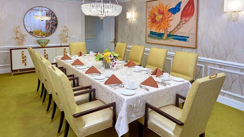 A formal dining room with a long rectangular table covered with a white patterned tablecloth. The table is set with white plates, cups, silverware, and brown folded napkins. There is a floral centerpiece in the middle of the table. Surrounding the table are ten yellow upholstered chairs with wooden armrests. The room has green carpet, patterned wallpaper, a round mirror on the wall, a sideboard with decorative items, a large colorful painting, and a chandelier hanging above the table.